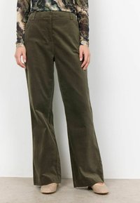 Pantaloni a gamba larga in corduroy verde oliva con una texture liscia, dotati di tasche frontali e vita sagomata. Indossati con scarpe beige con borchie.