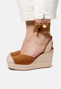 Hnědé semišové klínové sandály s tkanými jutovými podrážkami, které mají síťovanou panel a křížové šněrování kolem kotníku. Kulatá špička a plochý podpatek.