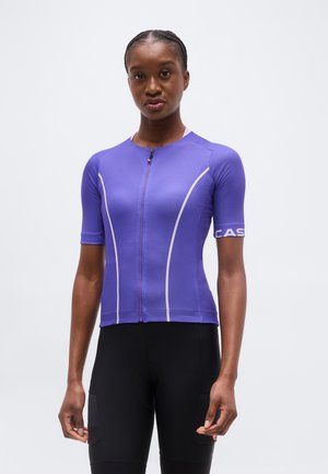 Sieviete valkā piegulošu violetas krāsas īsroku riteņbraukšanas kreklu ar priekšējo rāvējslēdzēju un melnus riteņbraukšanas šortus uz vienkārša fona.