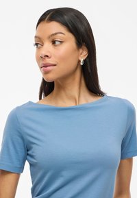 Blå bomullströja med korta ärmar och bred halsringning; slät textur. Modellen bär silverfärgade hängande örhängen. Vit bakgrund.