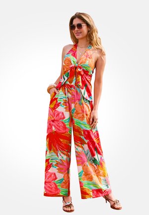 Femme portant un haut dos nu fleuri coloré et un pantalon large, des lunettes de soleil, des sandales et des bijoux, debout, les mains dans les poches.