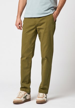 Homme portant un pantalon droit vert olive, une chemise bleu clair et des baskets blanches avec des détails beige et vert, debout avec une main dans la poche.