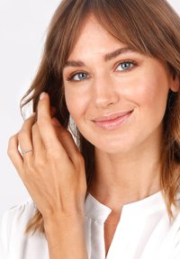 Silberner Ring mit einer kleinen Verzierung an der Hand einer Frau, die eine weiße Bluse trägt, umrahmt von lockigem braunem Haar und natürlichem Makeup.