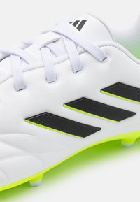 Botín de fútbol blanco hecho de material sintético con acentos en negro y verde neón, que presenta rayas texturizadas y cordones tradicionales.