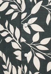 Zbliżenie na ciemną koszulę z tkaniny z białym wzorem liści i białymi guzikami wzdłuż środkowego szwu.