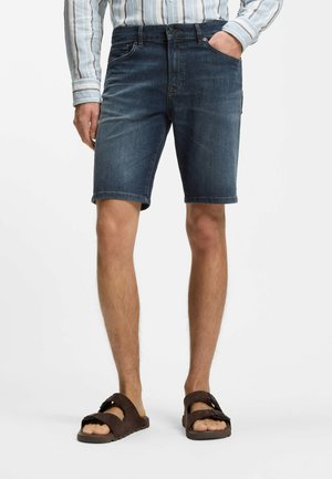 Mann trägt dunkelblaue Jeansshorts, gestreiftes Langarmshirt und braune Sandalen, steht mit einer Hand in der Tasche.