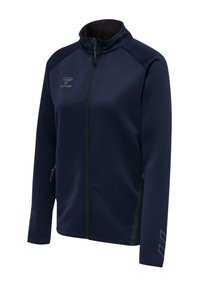 Marineblaue Sportjacke mit durchgehendem Reißverschluss, hohem Kragen, seitlichen Reißverschlusstaschen, Logo auf der Brust und dezenten Chevron-Details an den Ärmeln.