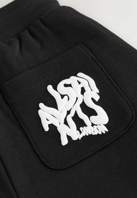 Melns auduma kabata uz apģērba ar baltu burbuļburtu tekstu "AllSaints London", kas iespiests uz kabatas.