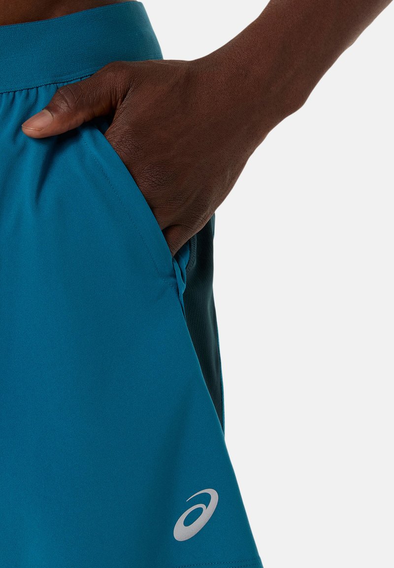 Shorts de sport turquoise avec une texture lisse, des poches latérales et un accent de logo sur l'ourlet inférieur gauche.