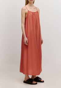 Langes, ärmelloses Kleid in einem sanften Korallton, mit gerafftem Ausschnitt und fließender Textur. Kombiniert mit schwarzen Sandalen mit gekreuzten Riemen.