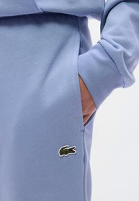 Ανοιχτό μπλε πουλόβερ με υφή υφάσματος και πλαϊνή τσέπη. Διαθέτει πράσινο κεντητό λογότυπο κροκόδειλου στο μπροστινό μέρος.