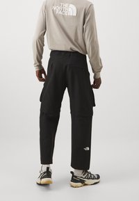 Czarne spodnie cargo z wieloma kieszeniami, elastycznym paskiem i logo na dolnej części nogawki, w połączeniu z butami trekkingowymi i koszulą z długim rękawem.