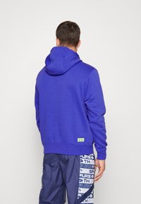 Felpa viola realizzata in materiale morbido con cappuccio regolabile, caratterizzata da una piccola toppa con logo verde vicino all'orlo. Abbinata a pantaloni corti scuri con fantasia.