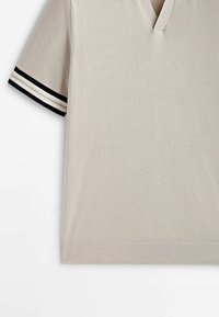Chemise beige à manches courtes avec un col en V et des accents rayés noirs et blancs sur les manches. Texture lisse avec un ourlet côtelé.