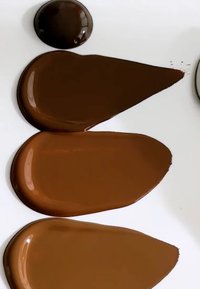 Drie kleurstaaltjes van bruine verf op een witte ondergrond: één cirkelvormig, twee langwerpige druppelvormen, met variërende tinten bruin en een glanzende textuur.