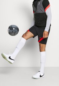 Chaqueta negra y gris de Nike con acentos rojos, combinada con pantalones cortos grises con una franja roja. Calcetas blancas y zapatos Nike, pateando un balón de fútbol negro.