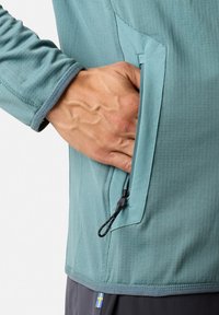 Hellblauer, strukturiert gewebter Pullover mit einem seitlichen Reißverschlussfach. Verstellbarer Kordelzug mit Stopper. Arm in der Position mit der Hand in der Tasche.
