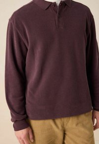 Mann in einem langärmligen, dunkelvioletten Fleece-Polohemd und hellbraunen Cordhosen, der vor einem schlichten beige Hintergrund steht.