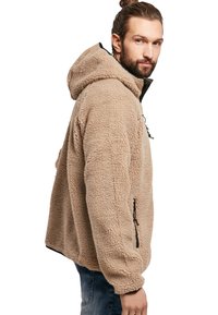 Chaqueta de forro polar beige con capucha, con una superficie texturizada, bolsillos con cremallera y detalles en negro a lo largo del frente y las mangas.