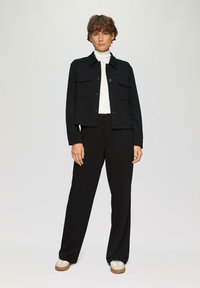 Veste noire courte avec deux poches avant, associée à un pantalon large noir et des baskets beiges. La tenue comprend un col roulé blanc en dessous.