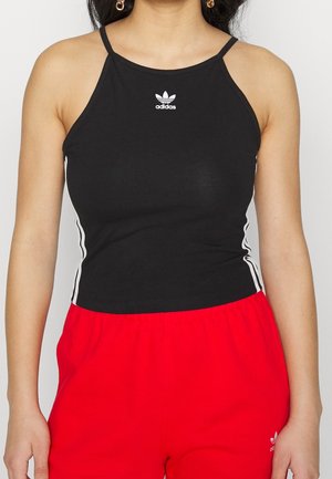 Kvinde iført sort Adidas ærmeløs top med hvide striber i siderne og røde Adidas bukser, stående mod en ensfarvet baggrund.