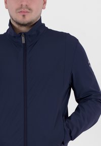 Giacca blu navy con zip e colletto alto. Realizzata in tessuto liscio e flessibile. Presenta un logo sottile sulla spalla sinistra.