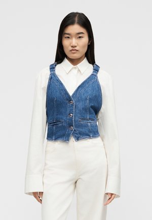 Kobieta ubrana w niebieską dżinsową kamizelkę zapinaną na guziki, na białej koszuli z długim rękawem i białych spodniach, stojąca na jednolitym tle.