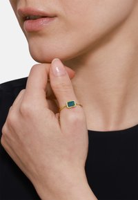 Goldring mit einem rechteckigen grünen Stein und einem schmalen Band. Die Hand ist in der Nähe des Kinns positioniert, vor einem neutralen Hintergrund.