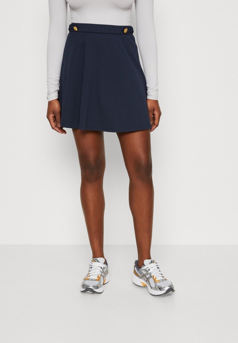 Personne portant une jupe bleu marine avec des boutons dorés, un haut blanc à manches longues et des chaussures de course argentées, blanches et orange, debout contre un mur blanc.