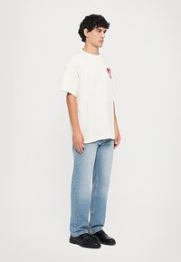 Cream short-sleeve t-shirt with a red heart logo on the chest, paired with light blue straight-leg jeans and black shoes. Minimalist design.