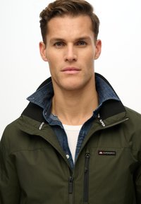 Chaqueta verde con cremallera y superficie texturizada, cuello negro y logo de Superdry. Debajo, se ve una camisa de mezclilla azul, con las capas abiertas.