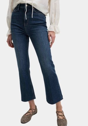 Femme portant un jean flare bleu foncé raccourci, un chemisier blanc à manches longues avec des poignets volantés, et des ballerines lacées imprimé léopard.