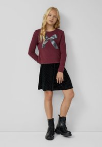 Bordowa koszula z długim rękawem z graficznym nadrukiem srebrnej kokardy w kropki, w połączeniu z czarną welurową spódnicą w białe kropki i czarnymi butami na sznurówki.