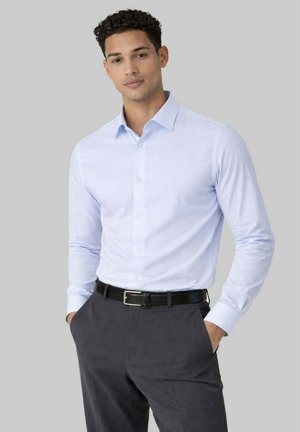 Homme portant une chemise bleue claire boutonnée, rentrée dans un pantalon foncé avec ceinture noire, mains dans les poches, debout devant un fond gris.