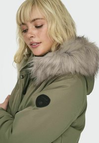Veste à capuche verte avec un col en fausse fourrure, dotée d'une finition texturée et d'un logo circulaire noir sur la manche.