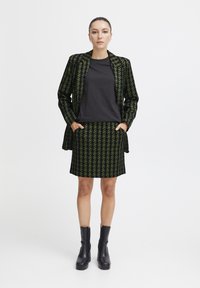 Zwarte t-shirt onder een groen en zwart houndstooth blazer en rok. Blazer heeft een getailleerde pasvorm; model draagt zwarte enkellaarsjes.