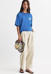 T-shirt en coton bleu oversized avec un design graphique, pantalon en lin beige clair et chapeau de paille multicolore. Sandales noires en plastique.