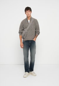 Suéter cinza de malha texturizada com fecho na frente, combinado com jeans azul escuro. O modelo usa uma bolsa tiracolo cinza e tênis brancos.