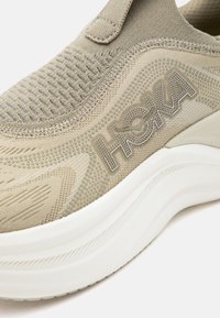 Μπεζ πλεκτό παπούτσι τύπου slip-on με παχύ λευκό πάτο και λογότυπο "HOKA" στο πλάι, με υφασμάτινες και ραμμένες λεπτομέρ ειες.