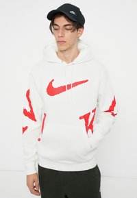 Bela jakna s kapuco z rdečim logotipom Nike, velikost oversized, spredaj ima žep. Oblečena z črnim kapo, kar ponuja sproščen stil športne obleke.