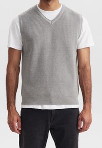 Grau gestrickte Weste mit V-Ausschnitt-Design, mit geripptem Muster und einfarbig, über einem weißen T-Shirt getragen, kombiniert mit dunklen Jeans.
