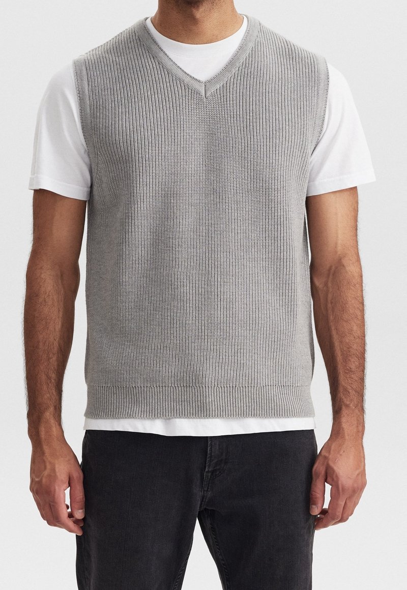 Grau gestrickte Weste mit V-Ausschnitt-Design, mit geripptem Muster und einfarbig, über einem weißen T-Shirt getragen, kombiniert mit dunklen Jeans.