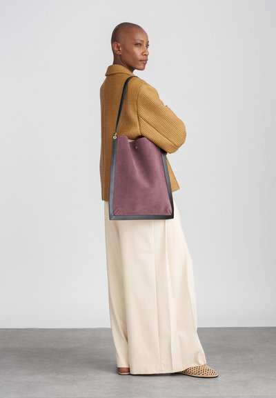 Paars suède schoudertas met zwarte leren accenten en gouden hardware, met een slouchy ontwerp. Draagt tegen een beige gebreide trui en crèmekleurige broek.