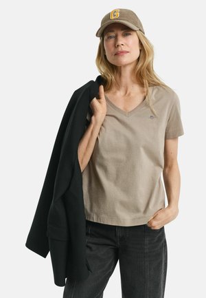 T-shirt beige à col en V en tissu doux, associé à un blazer noir. Le mannequin porte une casquette marron avec une lettre "G" jaune à l'avant.