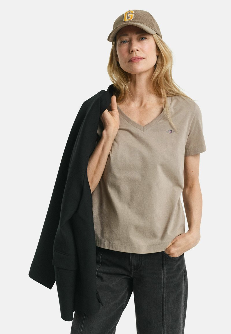 Beige V-hals t-shirt i blødt stof, parret med en sort blazer. Modellen har en brun kasket på med et gult bogstav "G" foran.