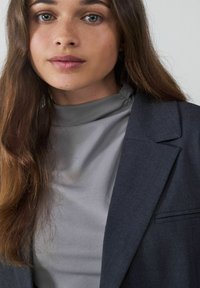 Blazer gris oscuro sobre un top de cuello alto gris claro, con texturas suaves y un ajuste entallado, y con el cabello largo y ondulado enmarcando el rostro.