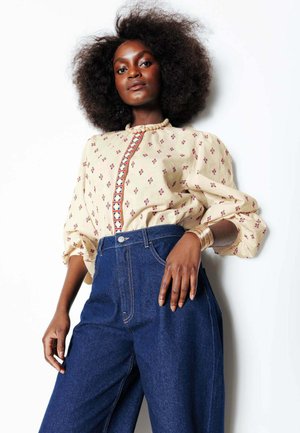 Femme noire aux grands cheveux bouclés, portant un chemisier crème à motifs, un jean taille haute bleu, des bracelets en or et un collier de perles.