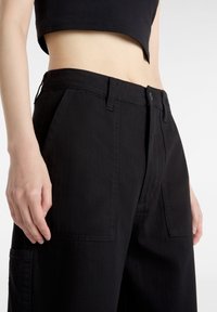 Svarta vida byxor i en bomullsblandning. Har framfickor, dragkedja med knappstängning och en texturerad yta.