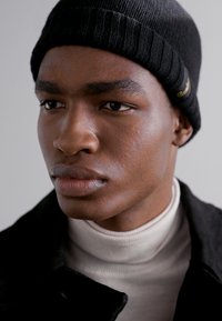 Gorro de malha canelada preto, usado com um suéter de gola alta bege claro. Close-up, destacando as feições faciais e a textura da pele contra um fundo neutro.
