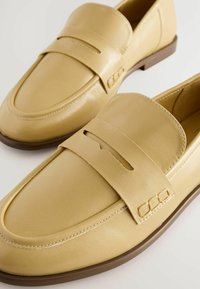 Light yellow leather loafers feature a smooth finish, a decorative strap, and a low brown rubber sole. The design includes stitching details.